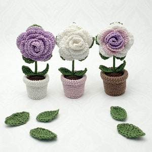 May include: Three crocheted flower arrangements in pots. The flowers are white, purple, and pink. Each flower has a green stem and leaves. The pots are white, pink, and brown.