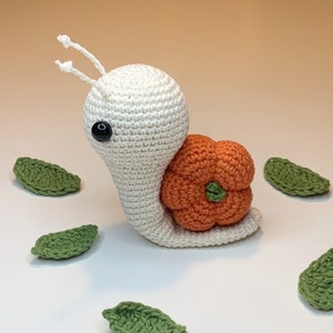 Puede incluir: Un caracol blanco de crochet con una gran flor naranja de crochet en su espalda. El caracol tiene ojos negros y dos antenas largas. Hay varias hojas verdes de crochet esparcidas alrededor del caracol.