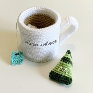 May include: A white crocheted mug with a brown crocheted interior, a green crocheted tea bag, and a green crocheted triangle with the letter "M" embroidered on it.