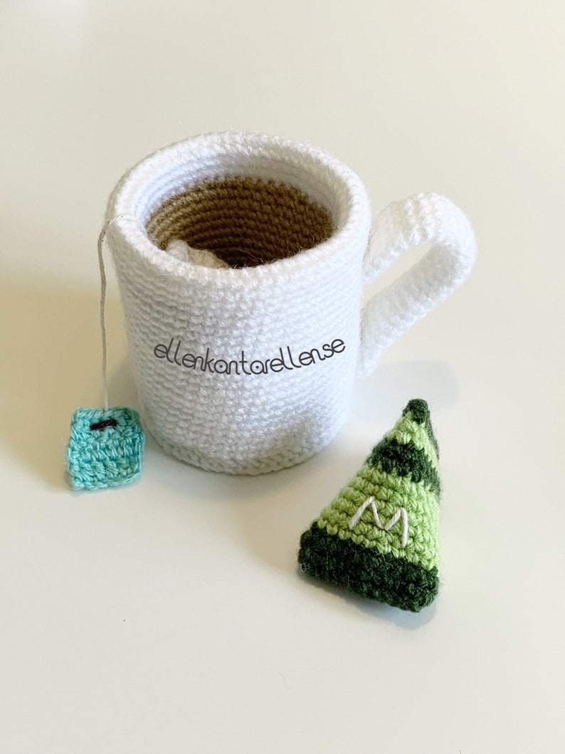May include: A white crocheted mug with a brown crocheted interior, a green crocheted tea bag, and a green crocheted triangle with the letter "M" embroidered on it.
