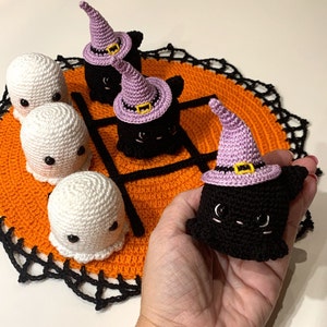 Puede incluir: Un juego de tic-tac-toe de Halloween tejido a crochet. Las piezas del juego incluyen fantasmas blancos y gatos negros con sombreros de bruja morados. El tablero es naranja con un borde negro.