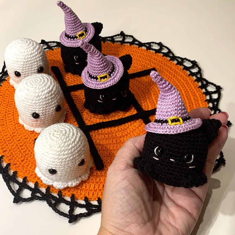 Puede incluir: Un juego de tic-tac-toe de Halloween tejido a crochet. Las piezas del juego incluyen fantasmas blancos y gatos negros con sombreros de bruja morados. El tablero es naranja con un borde negro.