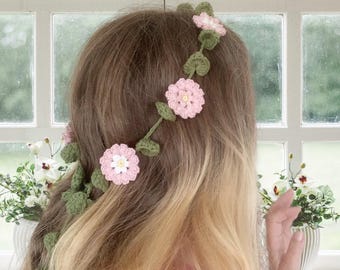 May include: A handmade crochet floral hair accessory featuring pink flowers with white centers and green leaves. The hairpiece is designed to be worn in the hair, adding a touch of nature-inspired charm. The flowers are arranged on a green vine.