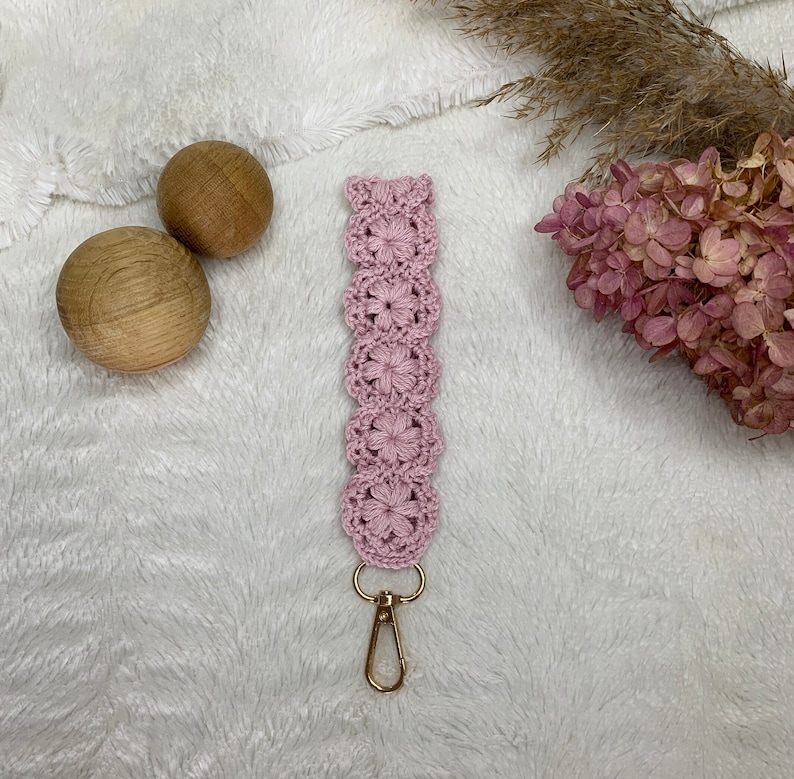 May include: A pink crocheted keychain with a gold metal clasp. The keychain is made of a delicate, floral pattern.