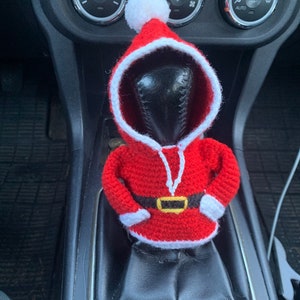 Puede incluir: Una figura de Pap&aacute; Noel de crochet en rojo y blanco, con un cintur&oacute;n negro, se coloca en la palanca de cambios de un coche.