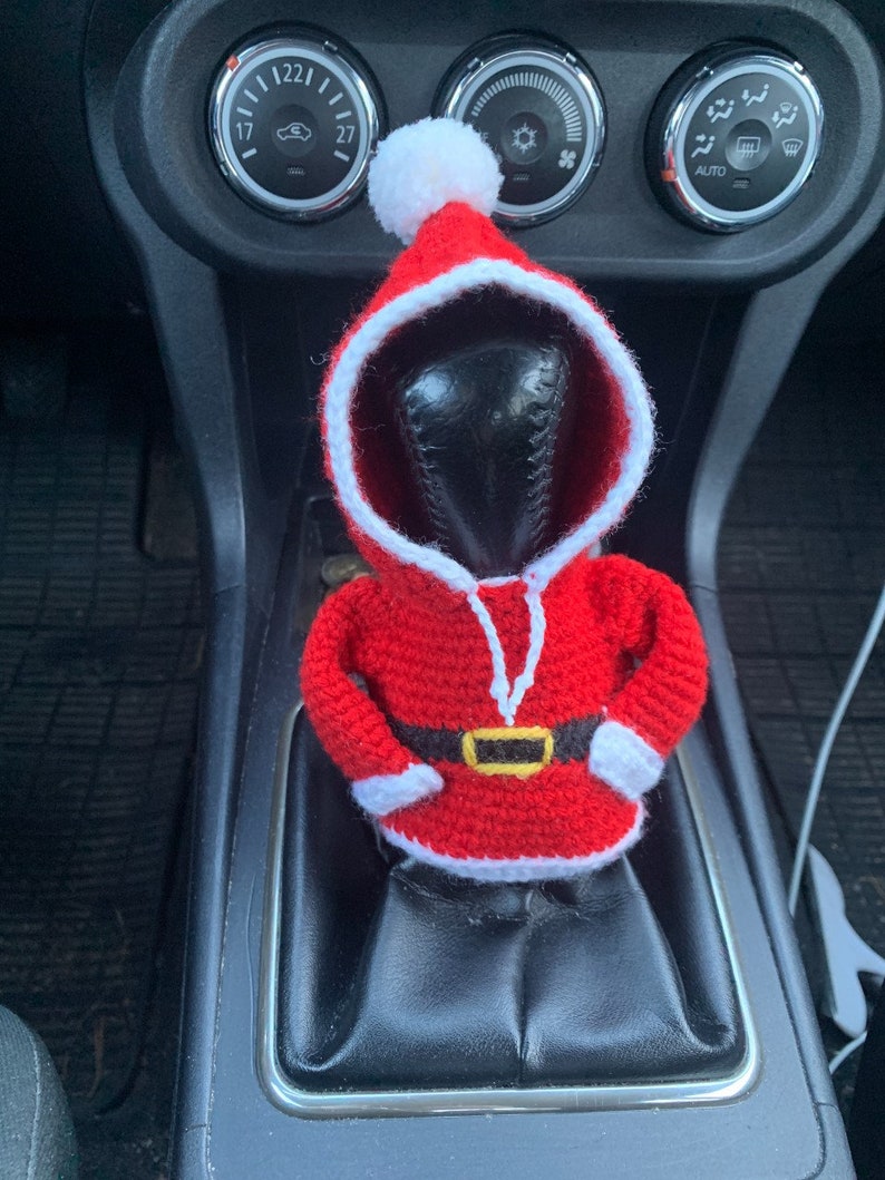 Puede incluir: Una figura de Pap&aacute; Noel de crochet en rojo y blanco, con un cintur&oacute;n negro, se coloca en la palanca de cambios de un coche.
