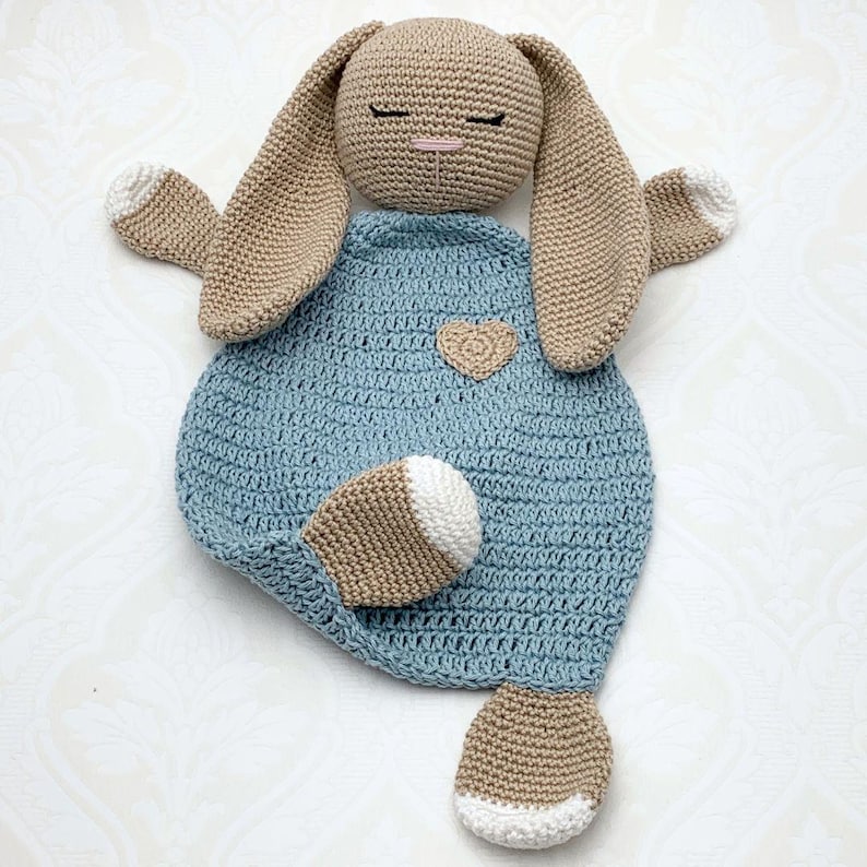 May include: A handmade crochet bunny lovey in tan and light blue. The bunny has long ears, closed eyes, and a small heart applique. The lovey is a soft, cuddly toy for babies and young children.