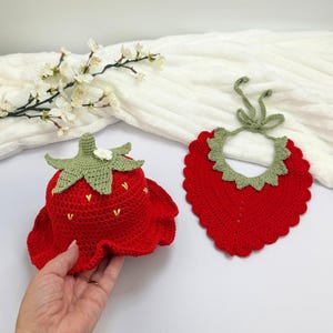 Könnte beinhalten: Ein roter Häkelhut in Erdbeerform mit grünem Stiel, einer kleinen weißen Blume und gelben Samen-Details, kombiniert mit einem passenden roten Häkellätzchen mit grünem Wellenrand und Bindebändern als Set.