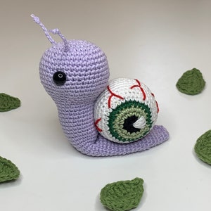 Puede incluir: Un caracol de ganchillo hecho a mano en lavanda con una gran concha de globo ocular. El globo ocular es blanco con venas rojas y un iris verde. Acentos de hojas verdes están dispersos.