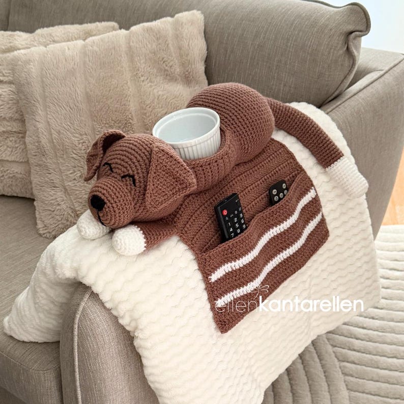 K&ouml;nnte beinhalten: Ein brauner, geh&auml;kelter Hunde-Couch-Organizer mit wei&szlig;em Schalenhalter, Taschen f&uuml;r Fernbedienungen und einem wei&szlig;-braunen Streifendesign. Der Hund hat wei&szlig;e Pfoten und eine wei&szlig;e Schwanzspitze. Der Organizer liegt auf einer wei&szlig;en Decke.