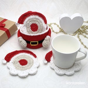 Puede incluir: Posavasos de ganchillo con temática de Papá Noel en rojo, blanco y beige. Un posavasos está debajo de una taza blanca. El diseño de Papá Noel incluye un gorro, barba y cinturón. Un corazón decorativo y una caja de regalo también están en la imagen. Decoración navideña.