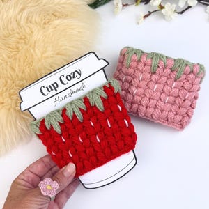 Peut inclure: Deux manchons de tasse au crochet avec un motif de fraise, corps en laine rouge et rose, feuilles en laine verte sur le dessus et accents en laine blanche. L'un est sur une carte blanche Cup Cozy Handmade, l'autre est posé à côté.