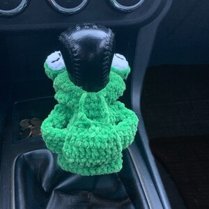 Pu&ograve; includere: Pomello del cambio per auto a forma di rana in crochet verde. La rana indossa una felpa con cappuccio e ha un pomello del cambio nero al centro.