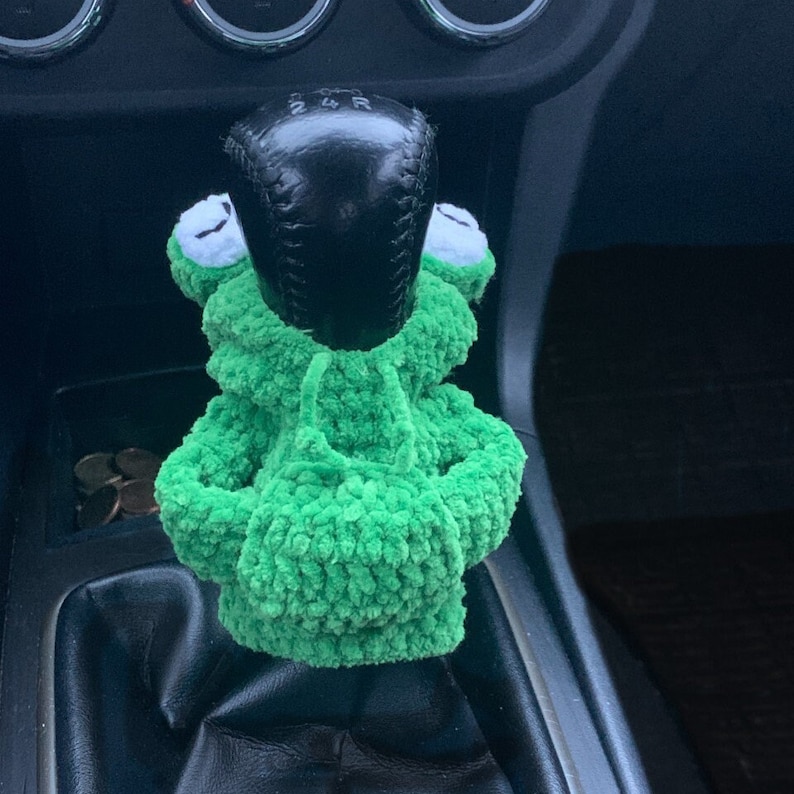 Pu&ograve; includere: Pomello del cambio per auto a forma di rana in crochet verde. La rana indossa una felpa con cappuccio e ha un pomello del cambio nero al centro.