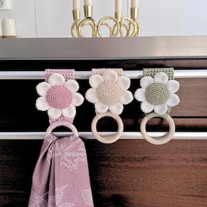 May include: Three crochet flower towel holders in pink, beige, and green, each with a wooden ring. A patterned towel hangs from the pink holder. The flowers have white petals and a contrasting centre. The holders are attached to a dark wooden surface.