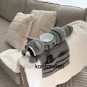 May include: A gray crochet raccoon-shaped sofa armrest organizer with a white bowl, two pockets, and a striped tail. The raccoon has black and white facial features. The organizer is resting on a sofa arm.