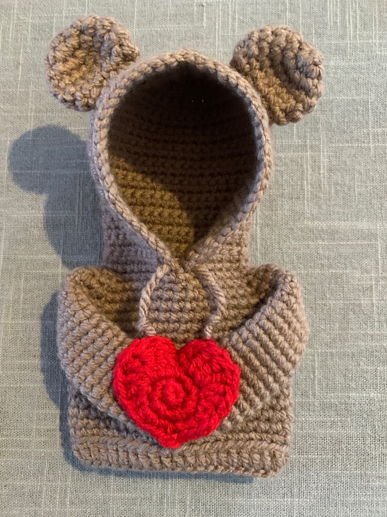 May include: A brown crocheted hooded sweater with bear ears and a red crocheted heart on the front.