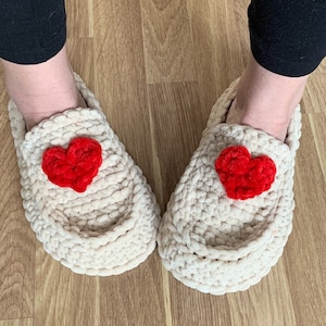 Op de afbeelding: Een paar crèmekleurige gehaakte pantoffels met rode hartversieringen. De pantoffels zijn gemaakt van zacht, pluche garen en hebben een comfortabele, ontspannen pasvorm.
