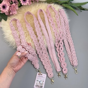 May include: A collection of pink crochet bag straps with gold-colored clasps. The straps feature various decorative patterns, including floral and openwork designs. The image also includes a matching crochet flower ring. The straps are displayed on a fluffy, off-white surface.