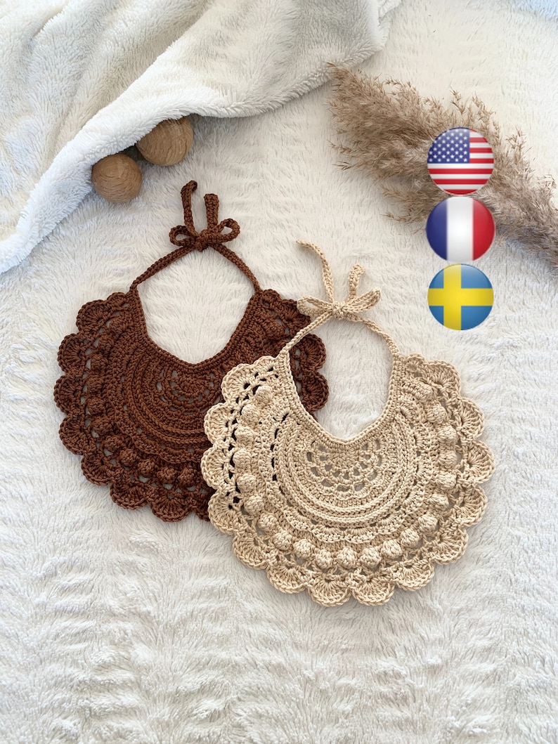 May include: Two crocheted baby bibs, one brown and one beige, with a scalloped edge and a row of small crocheted balls around the neckline. The bibs are laid out on a white fluffy surface.