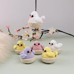 Op de afbeelding: Een verzameling handgemaakte gehaakte vogels in verschillende kleuren, waaronder wit, geel, roze en lavendel. Elke vogel zit in een beige mandje. Eén vogel hangt aan een zwart touwtje. De vogels hebben zwarte ogen en oranje snavels.