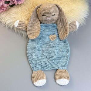 May include: A handmade crochet bunny lovey in beige and light blue. The bunny has long ears, closed eyes, and a small heart applique on the front. The bunny's feet and the tips of its hands are white. The lovey is designed for babies and young children.