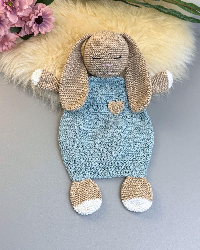 May include: A handmade crochet bunny lovey in beige and light blue. The bunny has long ears, closed eyes, and a small heart applique on the front. The bunny's feet and the tips of its hands are white. The lovey is designed for babies and young children.