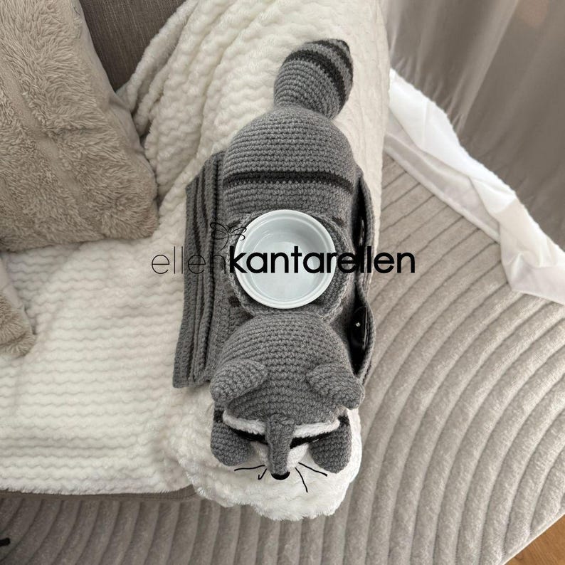 May include: A gray crocheted raccoon-shaped cup holder with a white bowl insert. The raccoon has a striped tail and a white face with black whiskers. The text "ellenkantarellen" is visible.