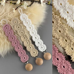 May include: Four crochet pacifier clips in pink, beige, and white, each with a wooden bead and metal clip. The clips are arranged on a light-colored, textured surface, with a sprig of white flowers in the background.