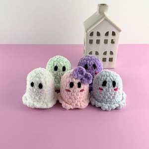 Puede incluir: Cinco figuras de fantasmas de ganchillo hechas a mano en blanco, verde claro, rosa, morado y azul claro. Cada fantasma tiene ojos negros y una boca sonriente. Una casa de cer&aacute;mica blanca con ventanas recortadas est&aacute; en el fondo. Los fantasmas est&aacute;n colocados sobre una superficie rosa.