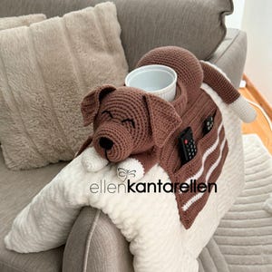 K&ouml;nnte beinhalten: Ein braun-wei&szlig; geh&auml;kelter Hunde-Sofa-Armlehnen-Organizer. Er hat einen Hundekopf, einen Becherhalter, Taschen f&uuml;r Fernbedienungen und den Text "ellenkantarellen". Der Hund liegt auf einer wei&szlig;en, flauschigen Basis.