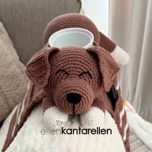 K&ouml;nnte beinhalten: Eine braun-wei&szlig; geh&auml;kelte Hunde-Decke mit einer wei&szlig;en Sch&uuml;ssel oben. Der Hund hat ein l&auml;chelndes Gesicht und wei&szlig;e Pfoten. Die Decke hat eine Tasche an der Seite. Der Text "ellenkantarellen" ist sichtbar.