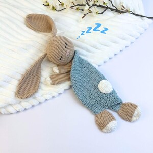May include: A crocheted bunny toy in shades of tan, blue, and white, lying on a white textured surface. The bunny has long ears, closed eyes, and a blue blanket. The word "zzz" is written above the bunny.