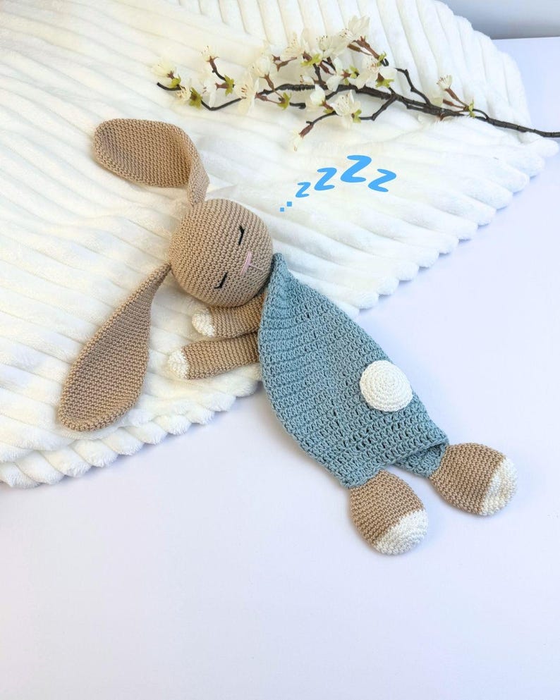 May include: A crocheted bunny toy in shades of tan, blue, and white, lying on a white textured surface. The bunny has long ears, closed eyes, and a blue blanket. The word "zzz" is written above the bunny.