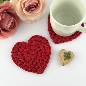 May include: Two red crocheted heart-shaped coasters on a white surface. One coaster has a white ceramic mug on top of it.