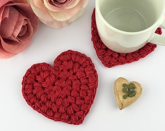 May include: Two red crocheted heart-shaped coasters on a white surface. One coaster has a white ceramic mug on top of it.