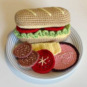 May include: A crocheted sandwich on a white plate with blue stripes. The sandwich is made of brown bread, green lettuce, red tomato, and yellow cheese. There are also two brown meat patties, a red tomato slice, a pink ham slice, and a yellow cheese slice on the plate.