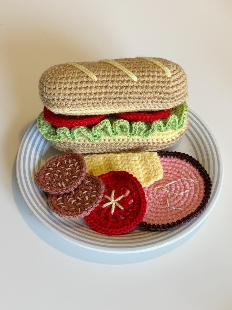May include: A crocheted sandwich on a white plate with blue stripes. The sandwich is made of brown bread, green lettuce, red tomato, and yellow cheese. There are also two brown meat patties, a red tomato slice, a pink ham slice, and a yellow cheese slice on the plate.