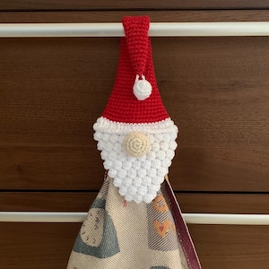 May include: A crocheted Santa Claus towel holder with a red hat and white beard. The holder is hanging on a white towel rack.