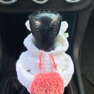 May include: White crocheted car gear shift cover with a pink pom-pom. The cover has a hoodie design with a white drawstring.