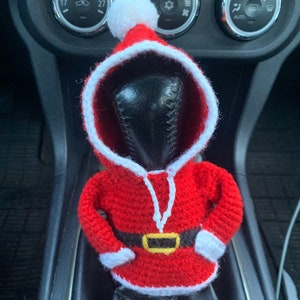Puede incluir: Una figura de Pap&aacute; Noel de crochet rojo y blanco con un cintur&oacute;n negro y un gorro de pomp&oacute;n blanco. La figura est&aacute; colocada en la palanca de cambios de un coche.