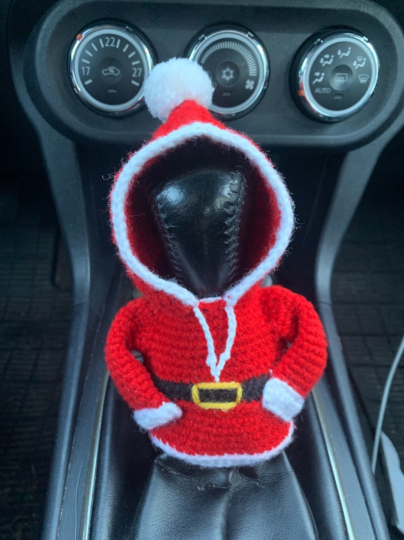 Puede incluir: Una figura de Pap&aacute; Noel de crochet rojo y blanco con un cintur&oacute;n negro y un gorro de pomp&oacute;n blanco. La figura est&aacute; colocada en la palanca de cambios de un coche.