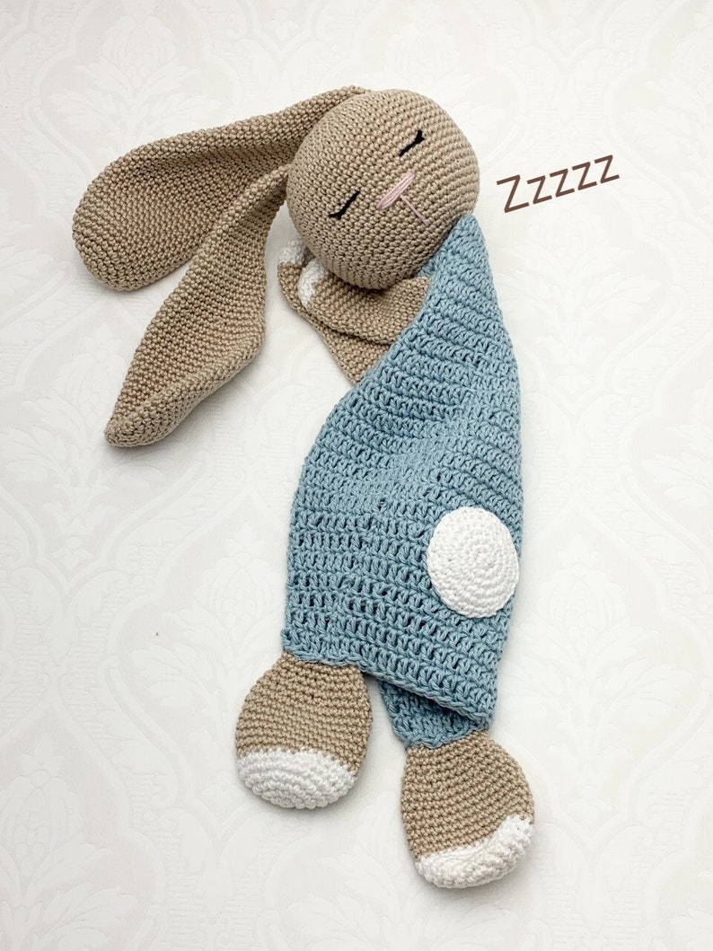 May include: A handmade crochet bunny in a sleeping pose. The bunny has long, floppy ears and is primarily tan with a light blue blanket. The blanket has a white circle detail. The word "Zzzzz" is written in brown.