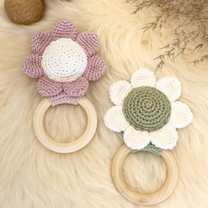 May include: Two crochet flower baby rattles. One has a pink flower with a white centre, the other has a white flower with a green centre. Both have a wooden ring handle.