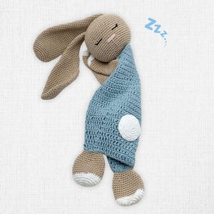 May include: A crocheted bunny toy in a sleeping pose. The bunny has long, floppy ears, a tan head and paws, and is wrapped in a light blue blanket. A white circle is on the blanket, and the word "Zzz..." is next to the bunny.