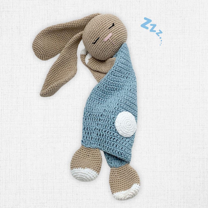 May include: A crocheted bunny toy in a sleeping pose. The bunny has long, floppy ears, a tan head and paws, and is wrapped in a light blue blanket. A white circle is on the blanket, and the word "Zzz..." is next to the bunny.