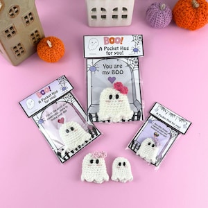Könnte beinhalten: Handgefertigte gehäkelte Geister-Taschenumarmungen, jeweils verpackt mit einer Karte mit der Aufschrift "Boo! A Pocket Hug for you!" und einer Nachricht. Die Geister haben schwarze Augen und einige haben rosa Schleifen. Halloween-Thema.