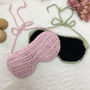 Könnte beinhalten: Zwei handgefertigte Schlafmasken aus Häkelarbeit. Eine ist hellrosa mit passendem Bindeband, die andere ist schwarz mit grüner Umrandung und Bindeband. Beide Masken sind aus weichem Garn gefertigt und sollen das Licht blockieren.