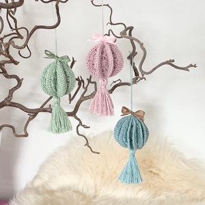 May include: Three crocheted ornaments with tassels hanging from a branch. The ornaments are pink, green, and blue.