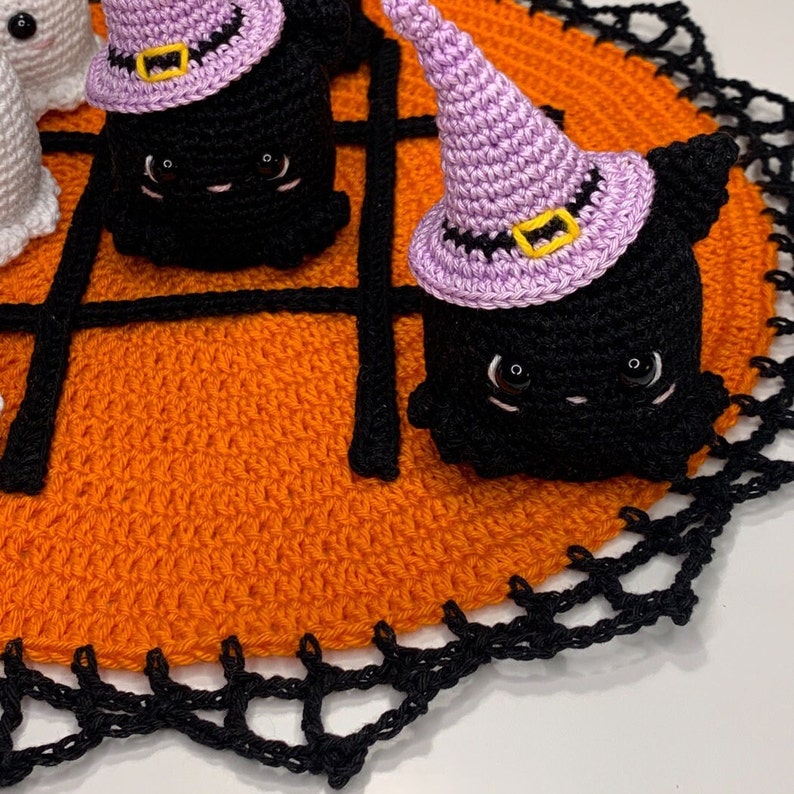 Puede incluir: Decoraciones de Halloween de ganchillo. Figuras de gato negro con sombreros de bruja morados con hebillas amarillas, colocadas sobre una alfombra naranja de ganchillo con un dise&ntilde;o de telara&ntilde;a negra. El fondo es blanco.